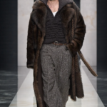 Modelo masculino en la pasarela de "The Portrait of Man", luciendo traje entallado en lana oscura con solapas amplias, reloj icónico transformado en broche sobre la solapa, camisa de cuello prominente abierta, pantalones cargo holgados.