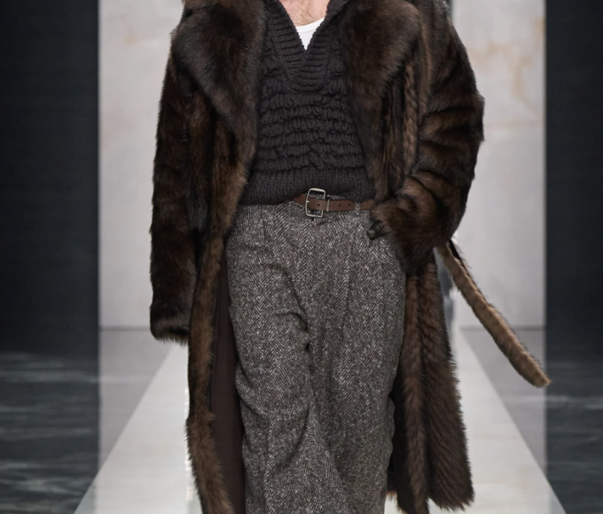 Modelo masculino en la pasarela de "The Portrait of Man", luciendo traje entallado en lana oscura con solapas amplias, reloj icónico transformado en broche sobre la solapa, camisa de cuello prominente abierta, pantalones cargo holgados.