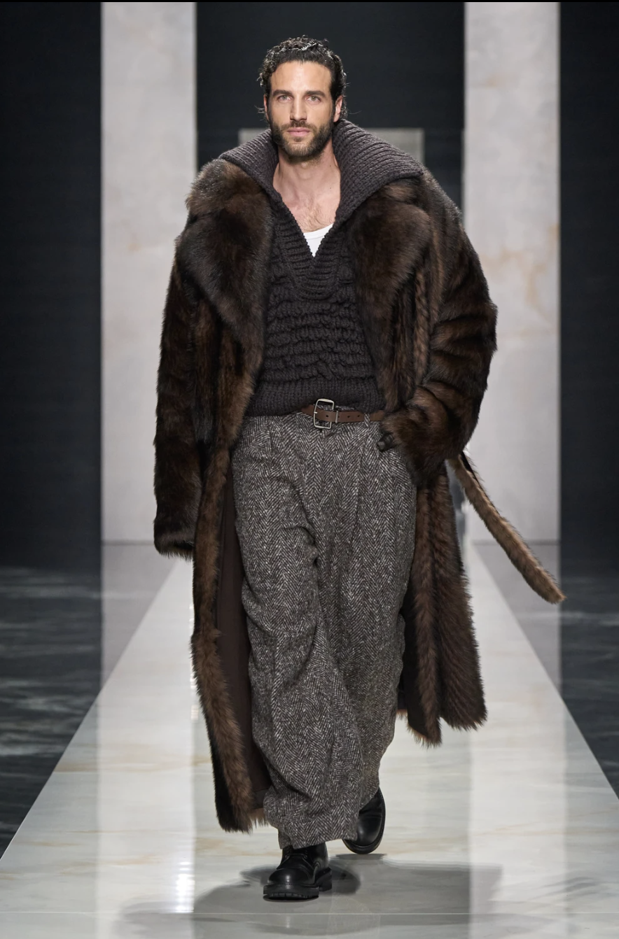 Modelo masculino en la pasarela de "The Portrait of Man", luciendo traje entallado en lana oscura con solapas amplias, reloj icónico transformado en broche sobre la solapa, camisa de cuello prominente abierta, pantalones cargo holgados.