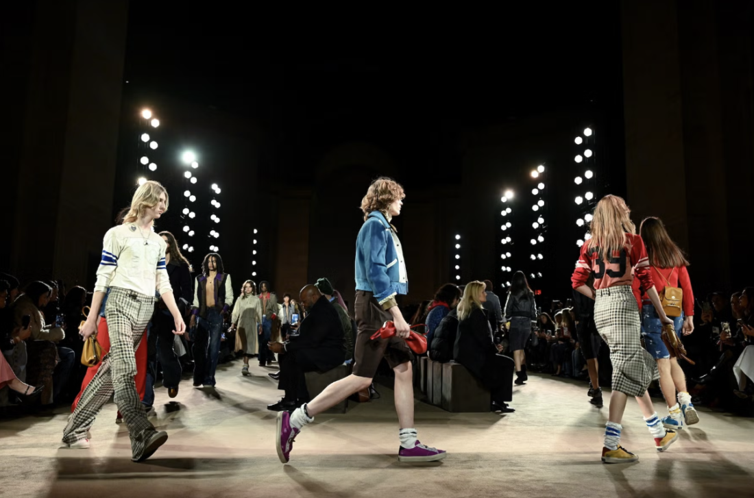 Modelo desfila en la pasarela de Coach NYFW con patchwork blazer entallado volteado mostrando forro estampado vibrante, Kisslock Frame Bag scrunched en cuero glove-tanned bajo el brazo.