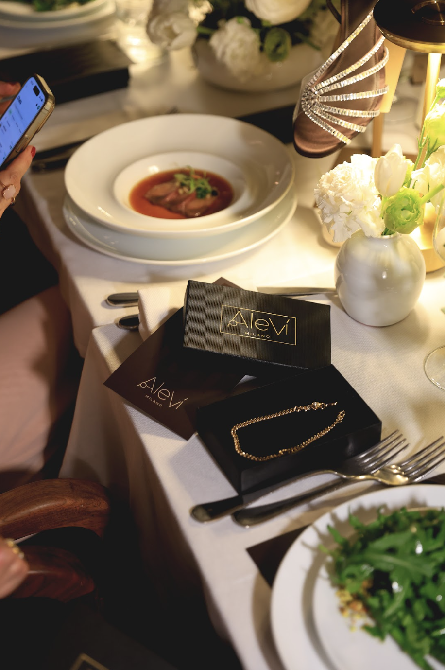 Grupo de invitadas elegantes en negro y nude alrededor de una mesa con velas en la cena de Aleví Milano FW26/27 en Blanco Castelar.