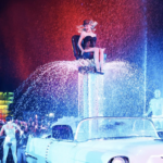 Sabrina Carpenter en el escenario principal de Coachella 2026, luciendo un minivestido Dior con flecos y brillo retro, rodeada de pantallas LED con efectos Hollywood, luces estroboscópicas y 100k fans eufóricos alzando celulares durante su hit "Espresso", capturando el vibe cinematográfico de su show histórico.