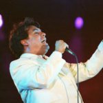 Juan Gabriel durante una presentación, vestido con uno de sus característicos trajes llamativos y ornamentados, reflejando el estilo teatral que marcó su trayectoria.