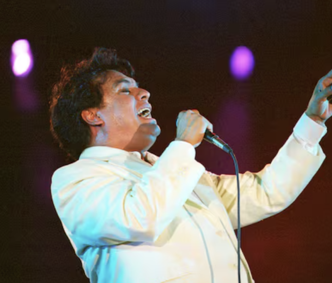 Juan Gabriel durante una presentación, vestido con uno de sus característicos trajes llamativos y ornamentados, reflejando el estilo teatral que marcó su trayectoria.