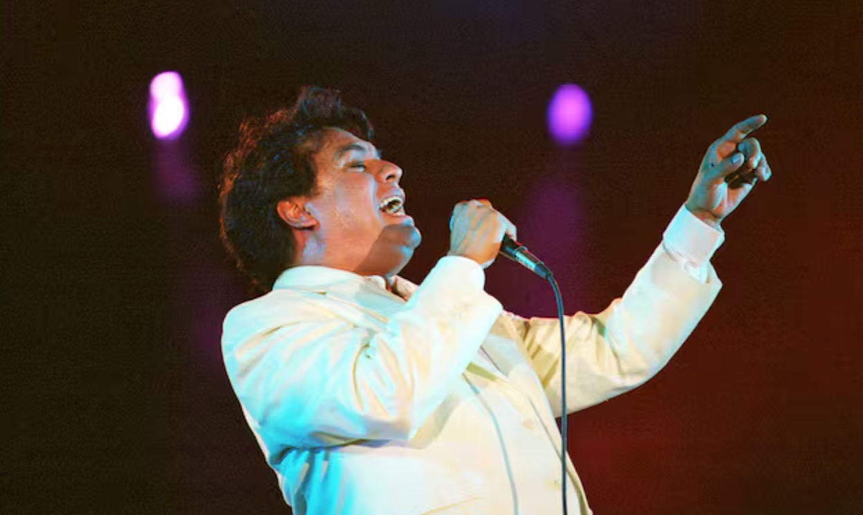 Juan Gabriel durante una presentación, vestido con uno de sus característicos trajes llamativos y ornamentados, reflejando el estilo teatral que marcó su trayectoria.